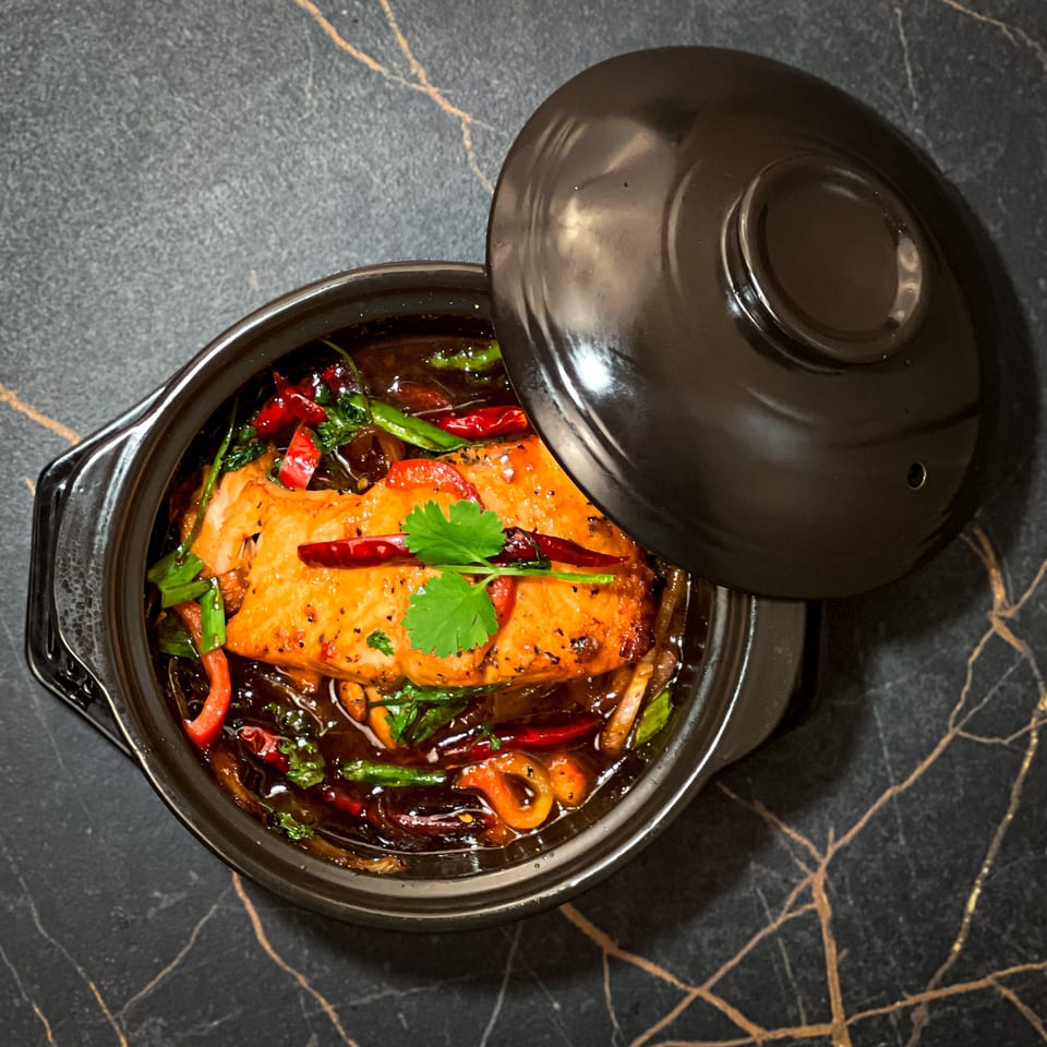 Braised salmon with red chilies and cilantro in a black ceramic pot on a dark marble surface.